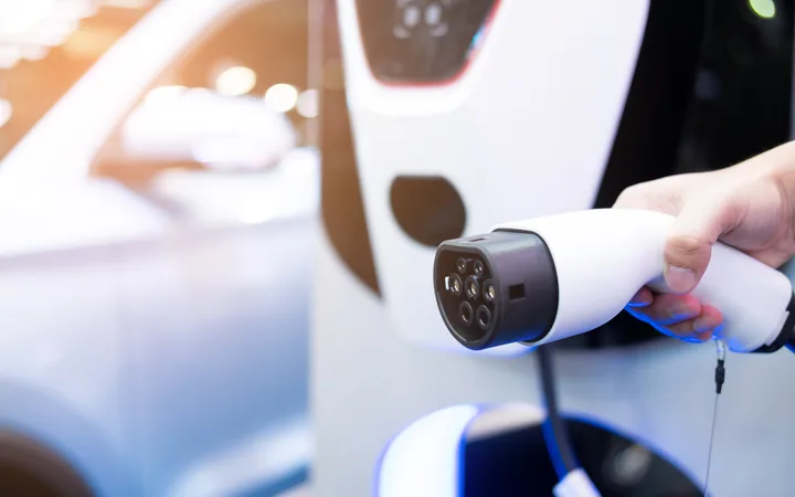 Close-up of a hand holding a white electric vehicle charging gun aimed at a car, with focus on the charging connector and port.