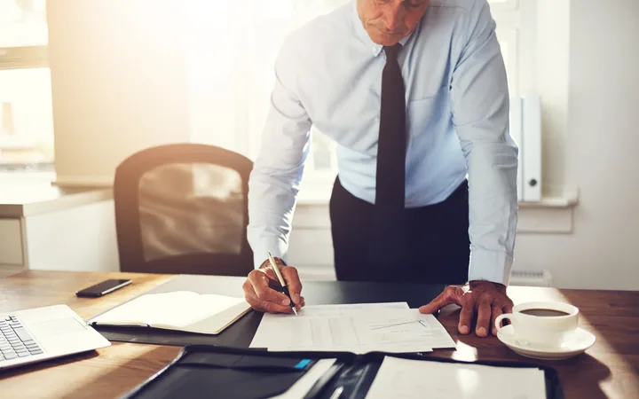 A man in a white shirt and black tie leans over a desk, writing on a document surrounded by office supplies, with sunlight streaming through windows.