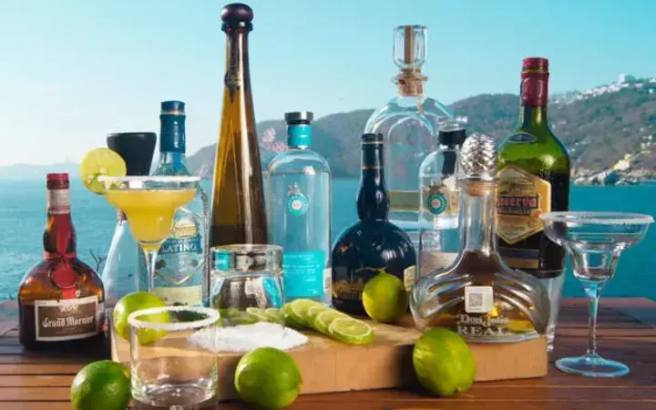 Assorted bottles of alcohol, glasses with drinks, and lime wedges arranged on a wooden surface with a seaside background and a hilly landscape.