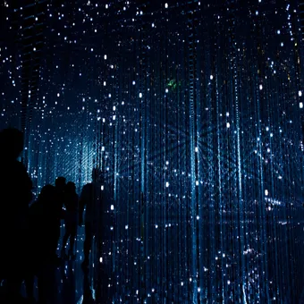 Silhouettes of people standing inside a dark, illuminated art installation with numerous hanging blue LED lights and reflective surfaces creating a starry effect.