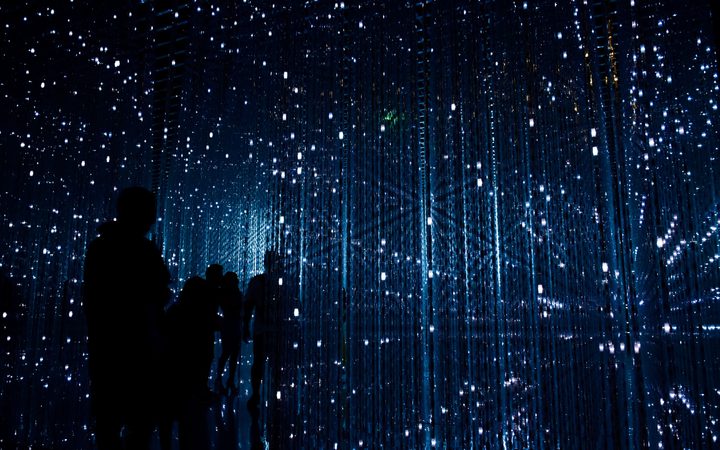 Silhouettes of people standing inside a dark, illuminated art installation with numerous hanging blue LED lights and reflective surfaces creating a starry effect.