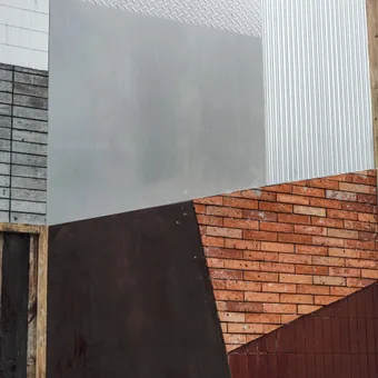 A close-up of various building materials including concrete blocks, metal, brick, wood, and painted surfaces arranged in a geometric pattern.