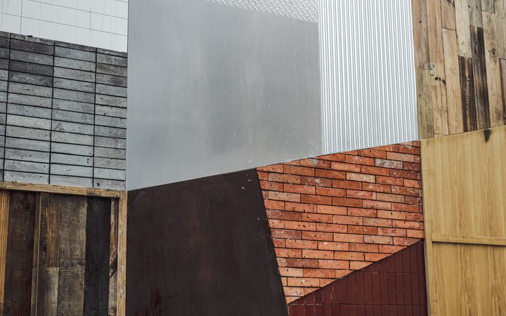 A close-up of various building materials including concrete blocks, metal, brick, wood, and painted surfaces arranged in a geometric pattern.