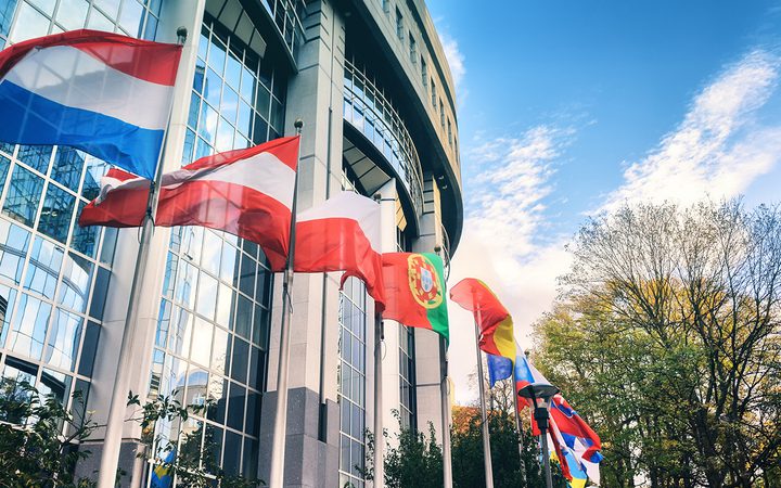 Flags of various countries flying outside a modern glass building on a partly cloudy day, with trees in the background and blue sky.