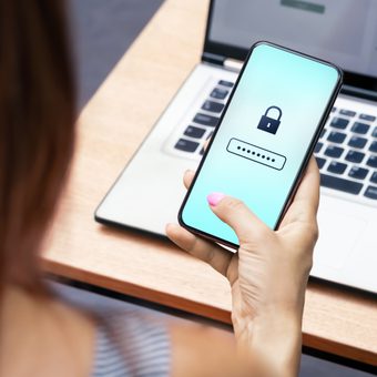 Person holding a smartphone displaying a padlock icon and a password field, with a laptop on a wooden desk in the background.