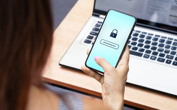 Person holding a smartphone displaying a padlock icon and a password field, with a laptop on a wooden desk in the background.