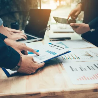 Group of people analyzing financial charts and data on a wooden table with laptops and smartphones during a meeting.