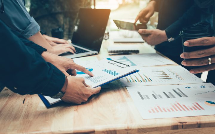 Group of people analyzing financial charts and data on a wooden table with laptops and smartphones during a meeting.