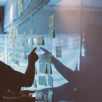 Person pointing at sticky notes on a glass wall during a meeting or brainstorming session, with light reflections and a window in the background.