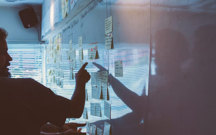Person pointing at sticky notes on a glass wall during a meeting or brainstorming session, with light reflections and a window in the background.
