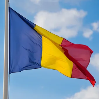 A French flag with blue, yellow, and red vertical stripes waving against a blue sky with white clouds.