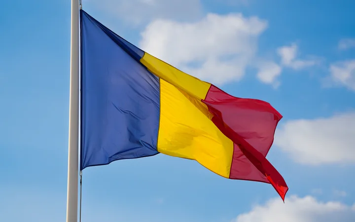A flag with blue, yellow, and red vertical stripes waving against a bright blue sky with scattered white clouds.