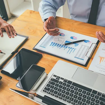 Two people discussing financial charts and data on a wooden table, surrounded by a laptop, tablets, a calculator, and documents with graphs.
