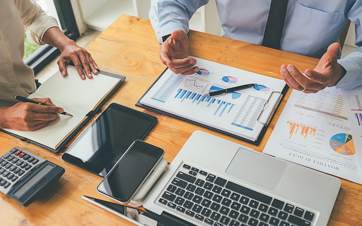 Two professionals are discussing business reports with charts and data on a wooden table, surrounded by laptops, tablets, and financial documents.