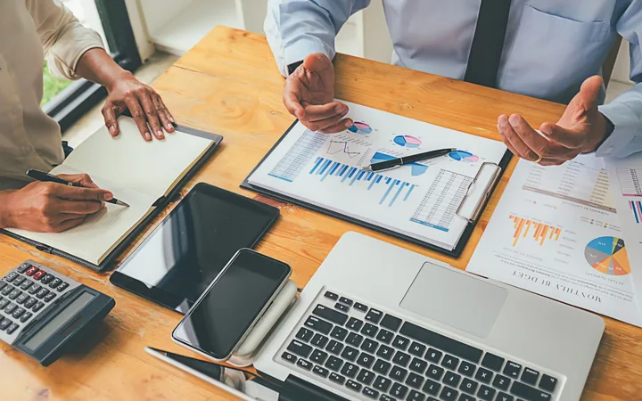 Two professionals are discussing business reports with charts and data on a wooden table, surrounded by laptops, tablets, and financial documents.