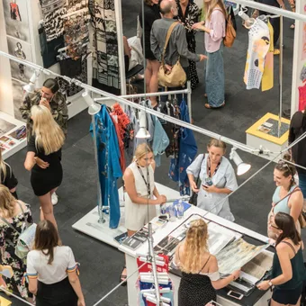 People browsing colorful art and fashion displays at an indoor exhibition or market stall. Some conversations, some examining artwork, vibrant decor.