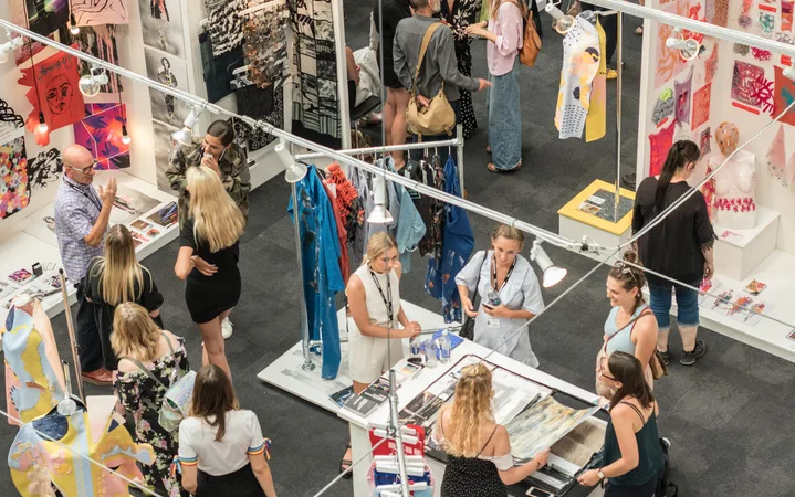 People browsing colorful art and fashion displays at an indoor exhibition or market stall. Some conversations, some examining artwork, vibrant decor.