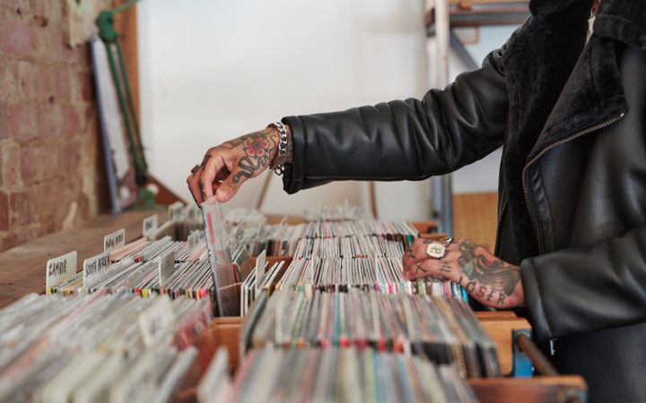 Tattooed person in a black leather jacket browses vinyl records in a brick-walled shop, reaching to pull a sleeve from a crate.