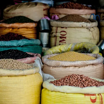 Bags of different grains and legumes are stacked in an open market, displaying a variety of colours, textures, and sizes.