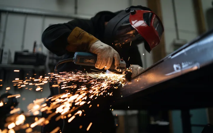 A worker wearing protective gear, including gloves and a helmet, welding metal with sparks flying in a workshop or garage setting.