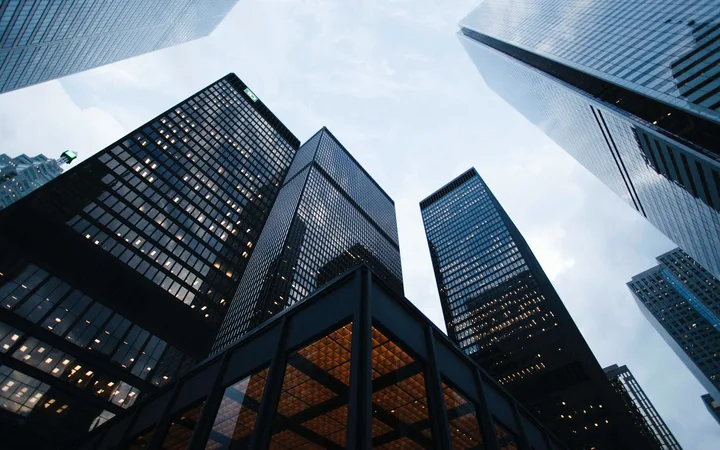 Tall modern skyscrapers viewed from below against a cloudy sky, reflecting light and creating a striking urban scene.