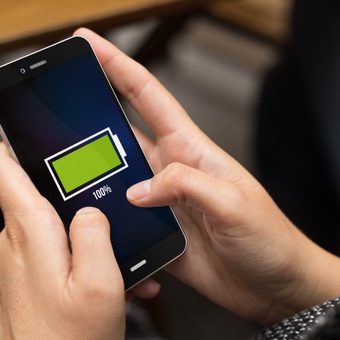 Person holding a smartphone displaying a full green battery icon at 100% on screen, with a blurred indoor background.