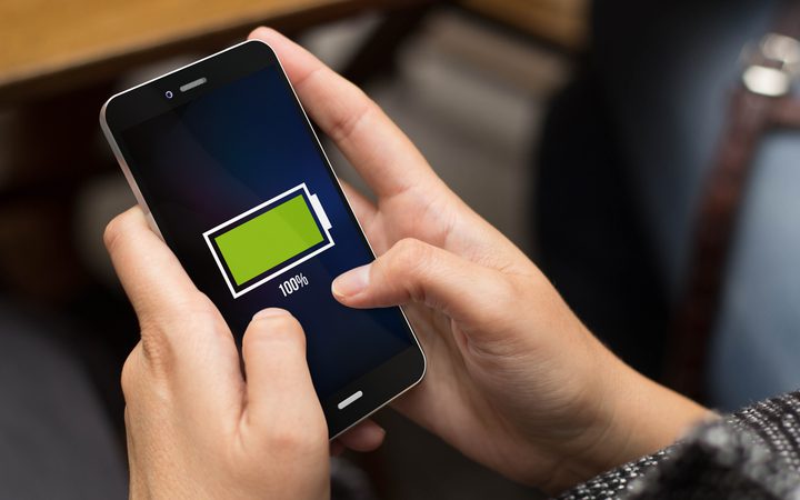 Person holding a smartphone displaying a full green battery icon at 100% on screen, with a blurred indoor background.