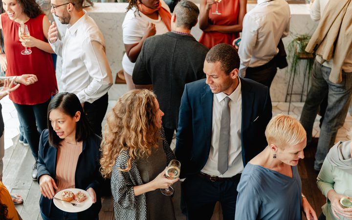 People socializing at a party, holding drinks, chatting, and mingling in a warmly lit indoor setting.