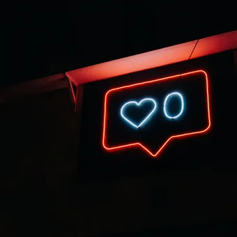 Neon sign resembling a social media "like" notification with red outline, white heart, and "O" in a speech bubble against a dark background.