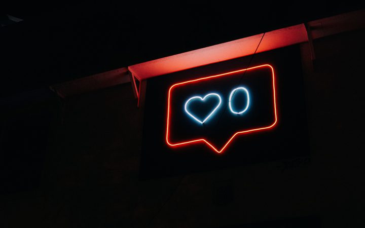 Neon sign resembling a social media "like" notification with red outline, white heart, and "O" in a speech bubble against a dark background.