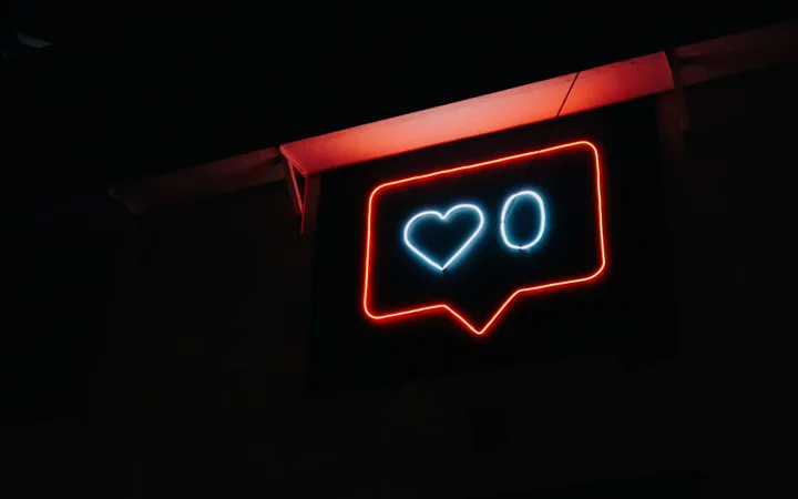 Neon sign resembling a social media "like" notification with red outline, white heart, and "O" in a speech bubble against a dark background.