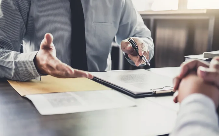 A person in business attire is gesturing while discussing documents during a meeting in a well-lit office.