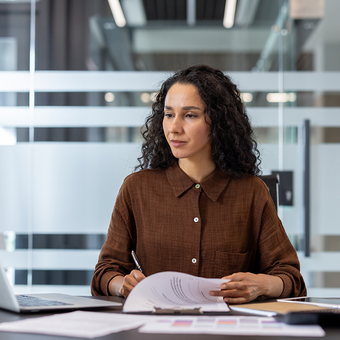 A person with curly dark hair sits at a desk in a modern office, reviewing papers with a pen beside an open laptop.