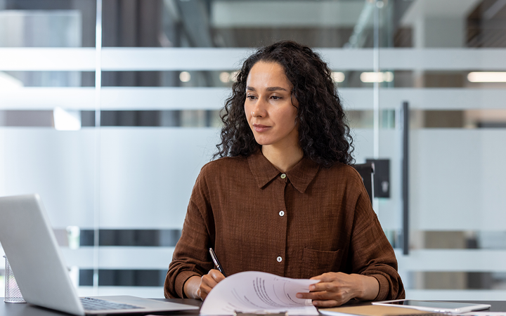 A person with curly dark hair sits at a desk in a modern office, reviewing papers with a pen beside an open laptop.