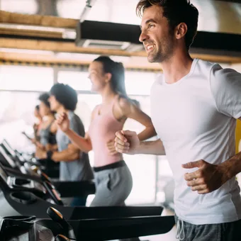 Group on treadmills in a bright gym, foreground person in a white T-shirt smiling as others run in the background.
