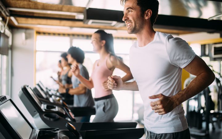 Group on treadmills in a bright gym, foreground person in a white T-shirt smiling as others run in the background.