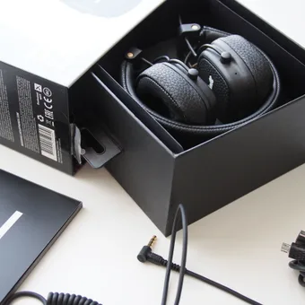 Black over-ear headphones inside a black box, with accessories including a user manual, audio cables, and a power adapter spread on a white surface.