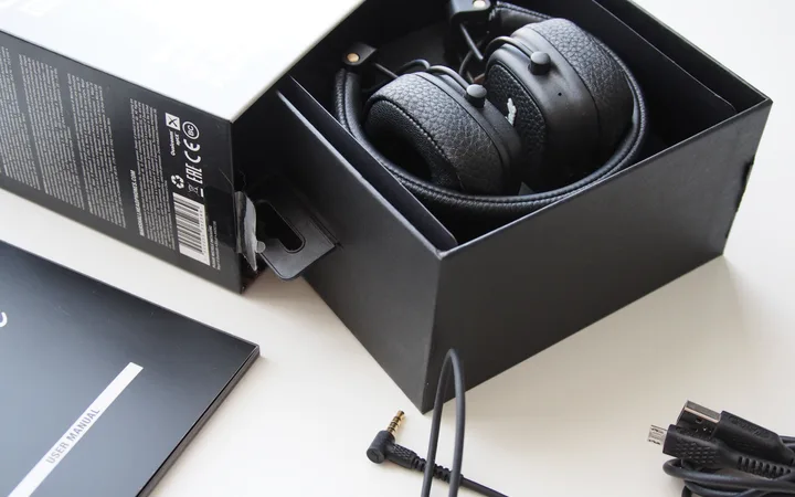 Black over-ear headphones inside a black box, with accessories including a user manual, audio cables, and a power adapter spread on a white surface.