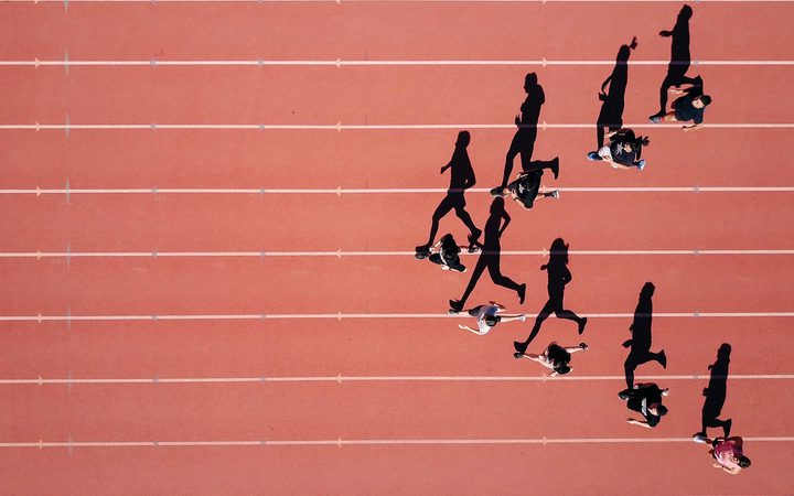 People jogging on a red outdoor track, casting long shadows, with white lane lines and a reddish pink background.
