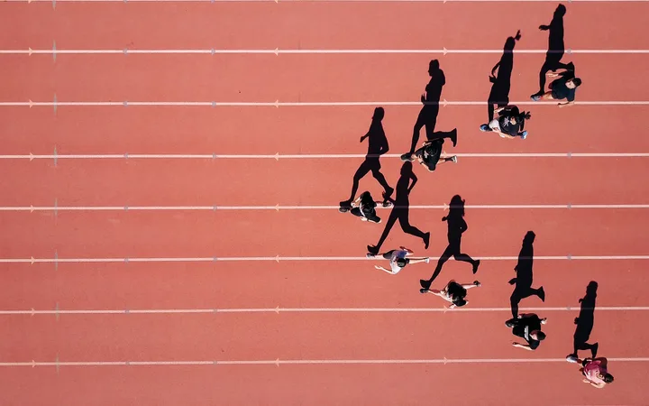 Bird's-eye view of people running and walking on a red track with white lane lines, casting shadows on the surface.