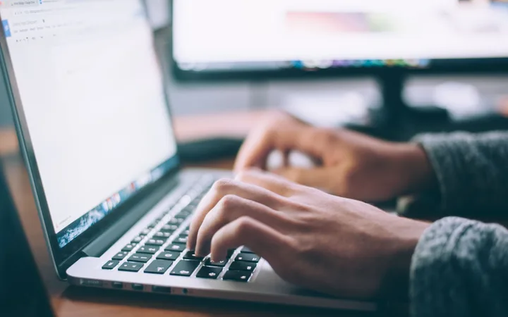Person typing on a laptop with dual monitors in the background, focusing on hands and keyboard in a workspace setting.