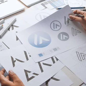 A hand holds a sheet with a blue circular logo, surrounded by design sketches, markers, rulers and a laptop on a cluttered desk.