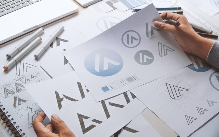 A hand holds a sheet with a blue circular logo, surrounded by design sketches, markers, rulers and a laptop on a cluttered desk.