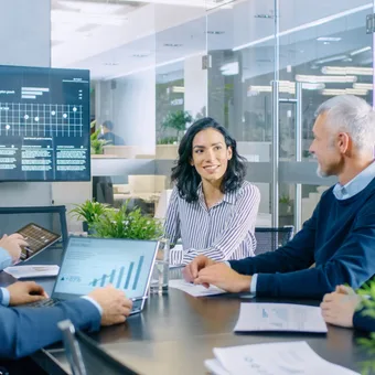 A diverse group of professionals in a modern conference room engaged in a discussion around a table, with a woman smiling and a presentation screen behind her.