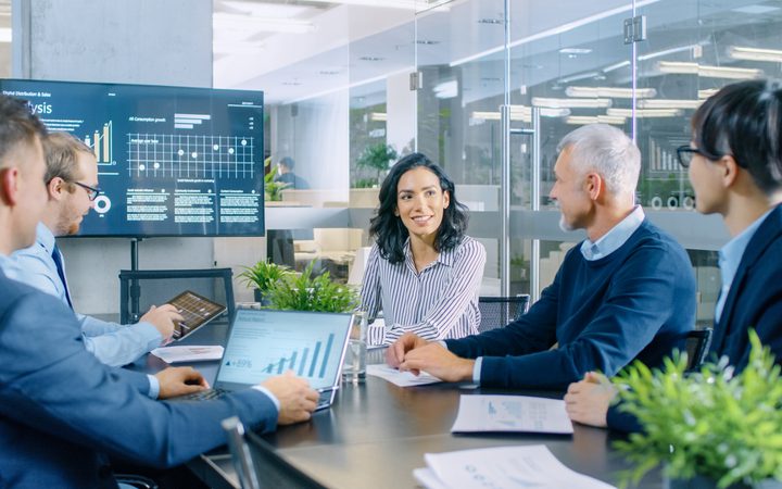 A diverse group of five professionals in a modern office meeting room engaged in a discussion, with a large digital screen displaying graphs and data behind them.