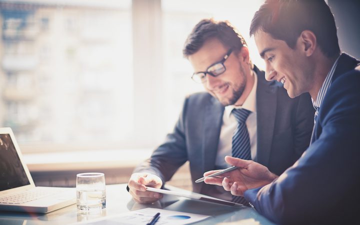 Two people in suits are discussing work at a table, smiling, with a laptop, a glass of water, and documents in a bright office.