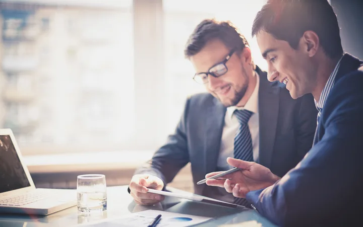 Two people in suits are discussing work at a table, smiling, with a laptop, a glass of water, and documents in a bright office.
