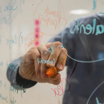 Person writing on a glass board with colorful markers, with handwritten notes and diagrams visible on the surface.