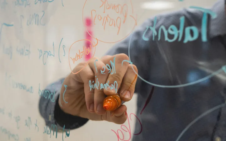 Person writing on a transparent board with colourful markers, holding an orange marker, surrounded by handwritten notes and diagrams.