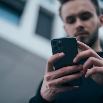 Person with dark hair and beard using a smartphone outdoors, with modern building architecture in the background.
